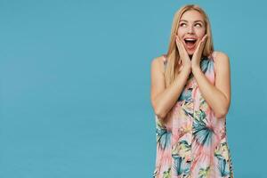 Indoor shot of pretty blonde female with long hair looking aside with wide mouth opened, keeping palms on her face while posing over blue background, showing her pleasant emotions photo