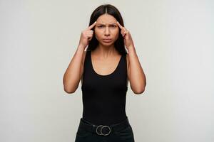 Portrait of displeased frowning young dark haired lady keepig forefingers on her temples while looking at camera with folded lips, suffering from headache, isolated over white background photo