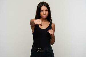 Indoor shot of young pretty dark haired woman with casual makeup boxing with raised fists and looking menacingly to camera, standing against white background photo