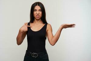 Studio shot of indignant young dark haired woman in casual black clothes pointing on herself with forefinger and frowning discontentedly her facce, isolated over white background photo