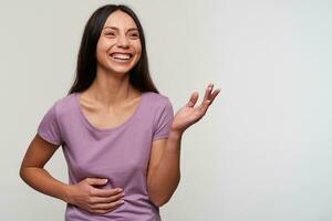 Joyful young lovely dark haired female with casual hairstyle holding on her belly while laughing happily, looking aside with wide cheerful smile, standing against white background photo