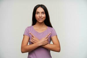 Open-eyed young pretty dark haired female with casual hairstyle crossing hands on her chest with raised forefingers, biting underlip while looking excitedly at camera, isolated over white background photo