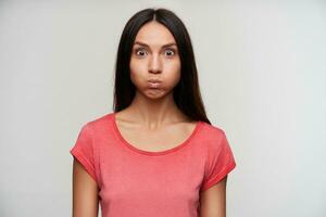 Indoor shot of open-eyed young brunette lady with natural makeup puffing out cheeks while looking surprisedly to camera, posing over white background with hands down photo