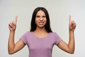 Displeased young brown-eyed brunette woman with natural makeup looking at camera with pout and frowning her face, posing over white background with raised forefingers photo