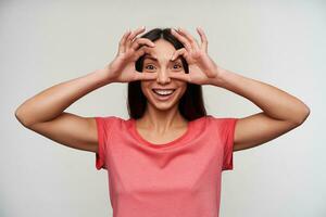 alegre joven atractivo de ojos marrones morena dama con natural maquillaje levantamiento manos a su cara y teniendo divertido mientras posando terminado blanco antecedentes en casual ropa foto