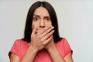 Frightened young brown-eyed dark haired female with casual hairstyle looking scaredly at camera and frowning her face, standing over white background with hands on her mouth photo