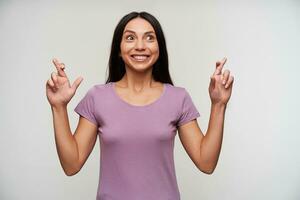 Cheerful young attractive brown-eyed lady in purple t-shirt looking happily aside with wide smile and raising hands with crossed fingers, standing against white background photo