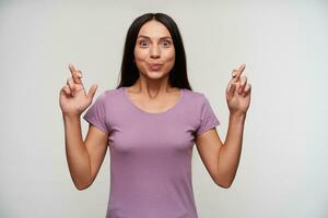 Excited young dark haired woman with casual hairstyle rounding eyes while looking joyfully at camera, raising hand with crossed fingers while posing over white background photo