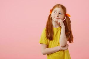 Portrait little pensive freckles red-haired girl with two tails, looks away, touches cheeks, wears in yellow t-shirt, stands over pink background. photo