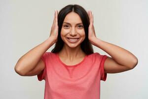 Cheerful young lovely dark haired lady with natural makeup closing her ears with hands and looking happily at camera and smiling widely, standing against white background photo