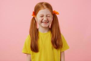 Portrait of little freckles red-haired girl with two tails, smiles with closed eyes, wears in yellow t-shirt, stands over pink background. photo