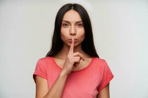 Portrait of charming young brunette woman raising forefinger in hush gesture and looking at camera with serious face, asking to be quiet while standing over white background photo