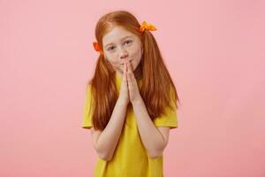 Portrait little sorry freckles red-haired girl with two tails, looks into the camera and cupped hands together, preyer gesture, wears in yellow t-shirt, stands over pink background. photo