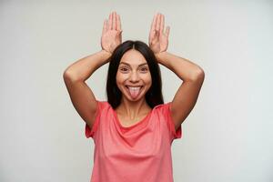 Funny photo of pretty young brown-eyed brunette female imitating rabbit ears with raised hands and showing happily her tongue to camera, posing over white background