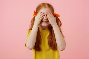 Photo of little freckles red-haired girl with two tails, smiles and covers eyes with palms, wears in yellow t-shirt, stands over pink background.