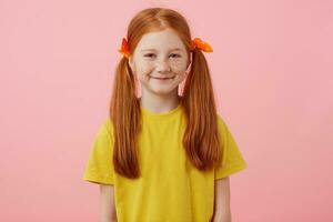 Portrait of petite freckles red-haired girl with two tails, looks into the camera and smiles, wears in yellow t-shirt, stands over pink background. photo