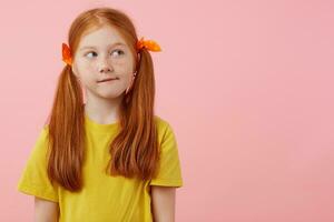 Photo of little thinking freckles red-haired girl with two tails, looks away, touches cheeks, wears in yellow t-shirt, stands over pink background with copy space.