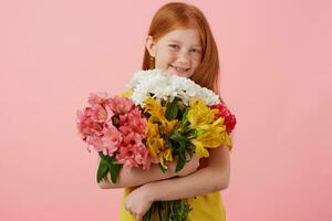 Portrait petite freckles red-haired girl with two tails, broadly smiling and looks cute, wears in yellow t-shirt, holds bouquet and stands over pink background. photo