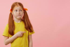 Little thinking freckles red-haired girl with two tails, looks away, wonts to draw you attentiot at the copy space and pointing finger to the right side, stands over pink background with copy space. photo