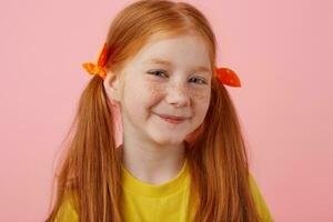 Close up of smiling petite freckles red-haired girl with two tails, smiling and looks cute, wears in yellow t-shirt, stands over pink background. photo