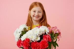 Happy petite freckles red-haired girl with two tails, broadly smiling and looks cute, wears in yellow t-shirt, holds bouquet and stands over pink background. photo