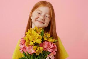 Portrait petite freckles red-haired girl with two tails, broadly smiling and looks cute, with closed eyes, holds bouquet, wears in yellow t-shirt, stands over pink background. photo