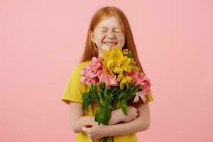 Petite freckles red-haired girl with two tails, with closed eyes broadly smiling and looks cute, holds bouquet, wears in yellow t-shirt, stands over pink background. photo