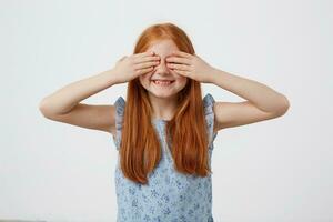 Photo of little freckles red-haired girl with two tails, smiles and covers eyes with palms, wears in blue dresst, stands over pink background.