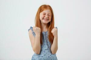 Portrait of happy smiling petite freckles red-haired girl, lwears in bluet, stands over white background with fists up. photo