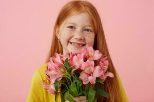 Portrait petite freckles red-haired girl with two tails, broadly smiling and looks cute, holds bouquet, wears in yellow t-shirt, stands over pink background. photo