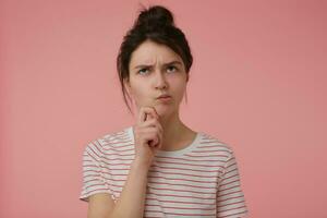 Young lady, pretty, serious looking woman with brunette hair and bun. Wearing t-shirt with red strips and wonder touching her chin. Emotional concept. Stand isolated over pastel pink background photo