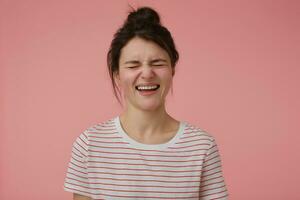 Portrait of attractive, joyful girl with brunette hair and bun. Wearing t-shirt with red strips and laughing with closed eyes. Emotional concept. Stand isolated over pastel pink background photo