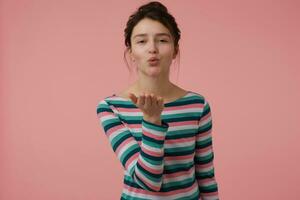 Teenage girl, happy looking, charming woman with brunette hair and bun. Wearing striped blouse and sending a kiss. Emotional concept. Watching at the camera isolated over pastel pink background photo