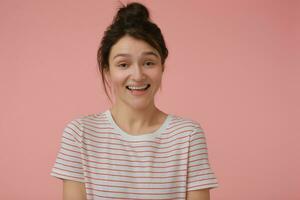 Young lady, pretty woman with brunette hair and bun. Wearing t-shirt with red strips and happy laughing. Emotional concept. Watching at the camera isolated over pastel pink background photo