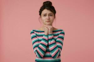 retrato de preguntando, triste mirando niña con morena pelo y bollo. vistiendo a rayas blusa y sostener manos debajo su mentón. emocional concepto. acecho a el cámara aislado terminado pastel rosado antecedentes foto