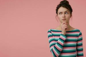 Teenage girl, curious looking woman with brunette hair and bun. Wearing striped blouse and touching her chin, eyebrow lifted. Watching to the left at copy space over pastel pink background photo