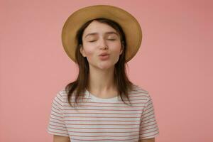 Nice looking woman, beautiful girl with long brunette hair. Wearing t-shirt with red strips and hat. Kissing you with closed eyes. Emotional concept. Stand isolated over pastel pink background photo