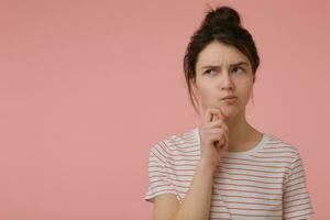 Portrait of attractive, wonder girl with brunette hair and bun. Wearing t-shirt with red strips and touching her chin. Emotional concept. Watching to the left at copy space over pastel pink background photo