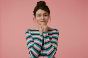 Teenage girl, happy looking woman with brunette hair and bun. Wearing striped blouse and biting her lip, asking. Emotional concept. Watching at the camera isolated over pastel pink background photo