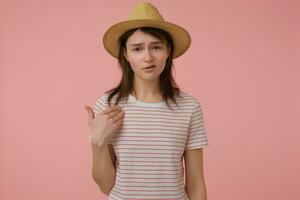 Serious looking woman, confident girl with long brunette hair. Wearing t-shirt with red strips and hat. Pointing at her self. Watching at the camera isolated over pastel pink background photo