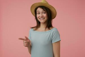 Nice looking woman, beautiful girl with long brunette hair. Wearing blueish t-shirt and hat. Watching at the camera and pointing to the left at copy space over pastel pink background photo