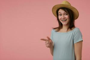 retrato de atractivo, adulto niña con largo morena cabello. vistiendo azulado camiseta y sombrero. acecho a el cámara y señalando a el izquierda a Copiar espacio terminado pastel rosado antecedentes foto