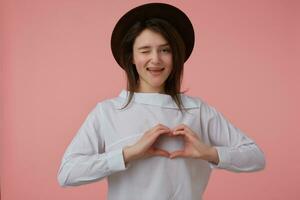 Portrait of attractive, adult girl with long brunette hair. Wearing white blouse and black hat. Showing love sign. Emotion concept. Watching and wink at the camera isolated over pastel pink background photo