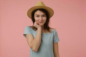 Nice looking woman, embarrassed girl with long brunette hair. Wearing blueish t-shirt and hat. Holding hand under her chin and looks flirty. Watching at the camera isolated over pastel pink background photo