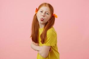 Portrait of petite freckles red-haired girl with two tails, looks and shows tounge at the camera, wears in yellow t-shirt, stands over pink background. photo