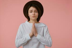 Teenage girl, begging looking woman with long brunette hair. Wearing white blouse and black hat. Holding palms together. Emotional concept. Watching at the camera isolated over pastel pink background photo