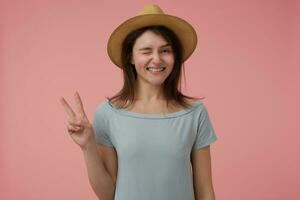 Portrait of attractive, adult girl with long brunette hair. Showing peace sign and wink smiling. Wearing blueish t-shirt and hat. Watching at the camera isolated over pastel pink background photo