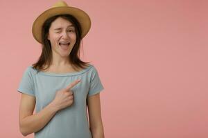 Portrait of attractive, adult girl with long brunette hair. Wearing blueish t-shirt and hat. Wink, smiling at the camera and pointing to the right at copy space over pastel pink background photo