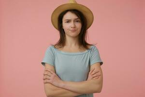 retrato de atractivo, adulto niña con largo morena cabello. vistiendo azulado camiseta y sombrero. pliegues su manos en un pecho. incrédulo acecho a el cámara aislado terminado pastel rosado antecedentes foto