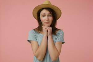 Nice looking woman, beautiful girl with long brunette hair. Wearing blueish t-shirt and hat. Look at you and beg for something. Watching at the camera isolated over pastel pink background photo